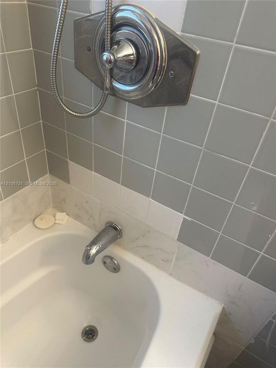 Bathtub featuring a stone-finish surround, chrome-finished wall-mounted faucet, and hand-held shower head with an integrated polished chrome pressure-balance valve on a square tile-accented wall