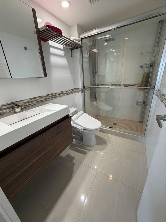 Modern bathroom featuring a wood-finish floating vanity with a white integrated sink, walk-in shower with a glass enclosure, decorative mosaic tile wall accent, and polished large-format floor tiling