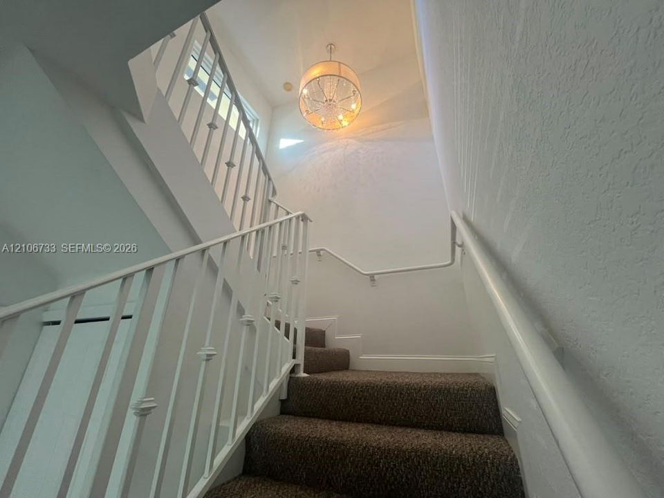 Staircase featuring a white metal balustrade, wall-mounted handrail, carpeted treads, and a multi-arm drum chandelier