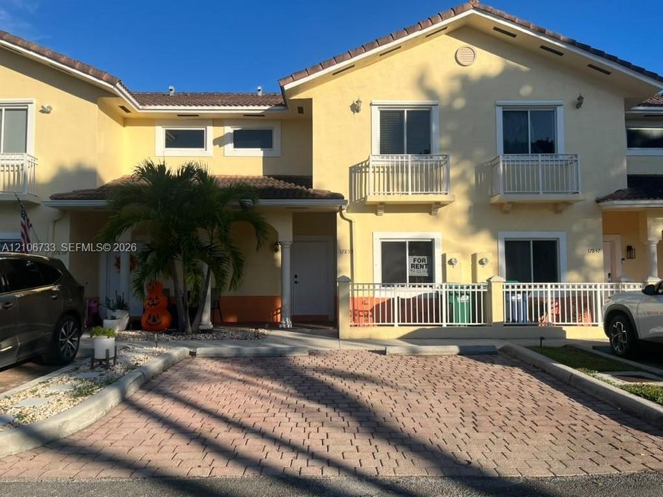 Two-story stucco townhouse exterior featuring a tiled roof, paver driveway, second-level balconies with metal railings, and a fenced front patio area