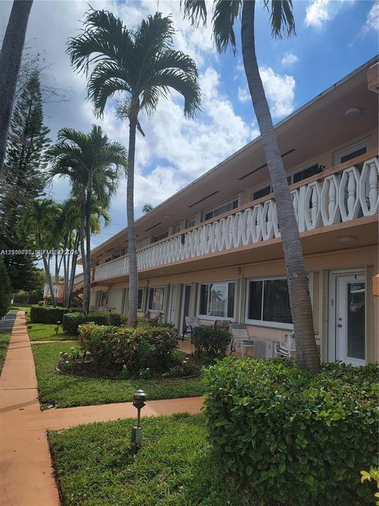 Two-story stucco building exterior featuring a geometric patterned balcony railing, mature palm trees, landscaped shrubbery, and paved pedestrian walkways