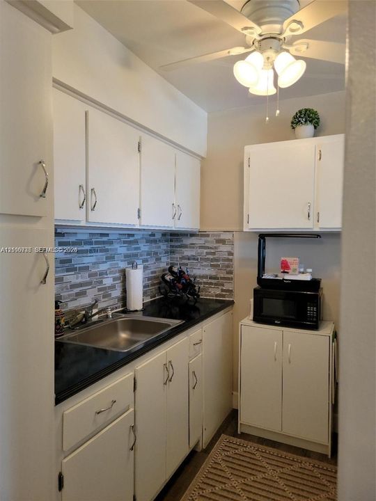 Kitchenette featuring white cabinetry, dark stone-finish countertops, stainless steel sink, multi-tonal linear tile backsplash, and a ceiling-mounted light fixture