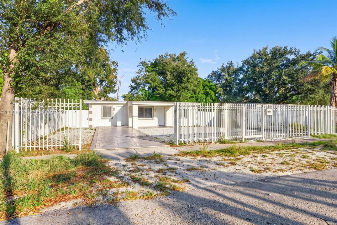Single-story residence featuring a paved concrete driveway, white vertical metal perimeter fencing, and mature trees