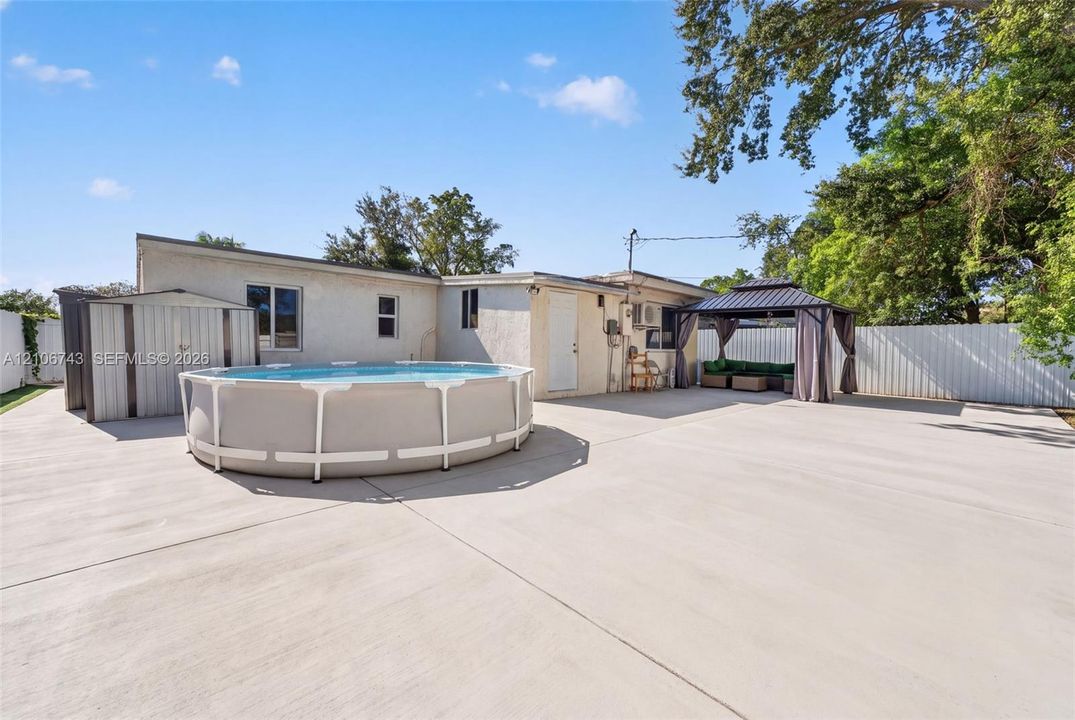 Expansive concrete patio featuring a permanent metal storage shed, above-ground pool, and built-in metal-frame gazebo enclosed by perimeter fencing