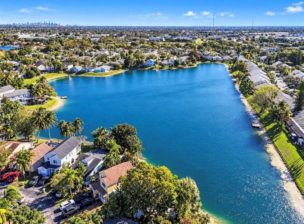 Aerial perspective of a lakefront residential community featuring expansive water frontage, mature tropical landscaping, and clear lines of sight to a distant cityscape skyline