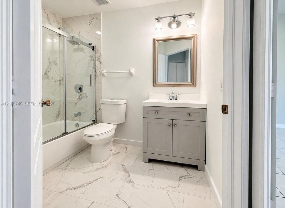 Bathroom featuring large-format veined tile flooring, a grey shaker-style vanity with a solid surface countertop, and a shower-tub combination with sliding glass doors and wall-to-ceiling tile surround