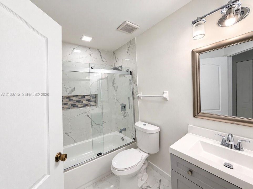 Bathroom featuring a grey vanity with an integrated rectangular sink, stone-patterned tile flooring, a shower-tub combination with glass sliding doors, stone-patterned wall tile, and a recessed decorative tile accent niche