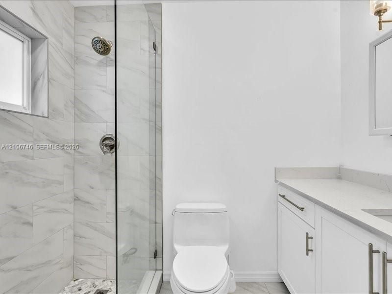 Walk-in shower with frameless glass enclosure, floor-to-ceiling stone-tile surround, vanity with stone-composite countertop, and wall-mounted light fixture