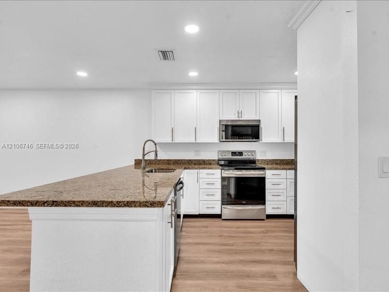 Kitchen featuring white shaker-style cabinetry, stone countertops, stainless steel appliances, a center island with a stainless steel sink, and wood-finish flooring