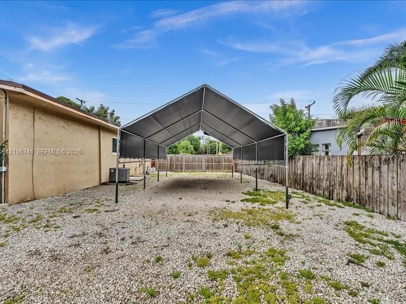 Metal-framed carport with a pitched canopy, positioned over a gravel surface and enclosed by a wooden privacy fence