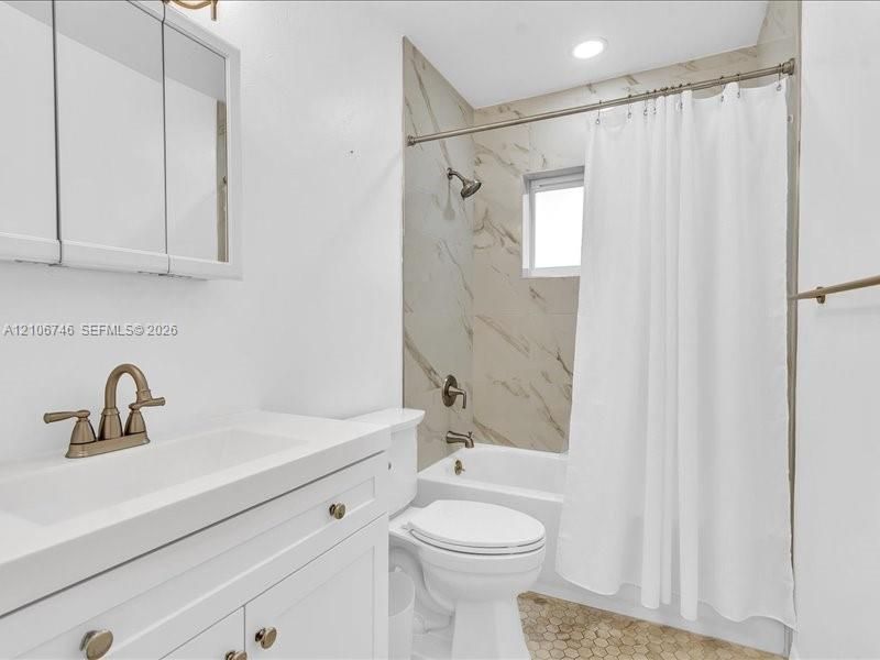 Bathroom featuring a white vanity cabinet, rectangular undermount sink with a bronze-finish faucet, recessed lighting, tub-shower combination with floor-to-ceiling stone-pattern tile surround, and hexagonal tile flooring