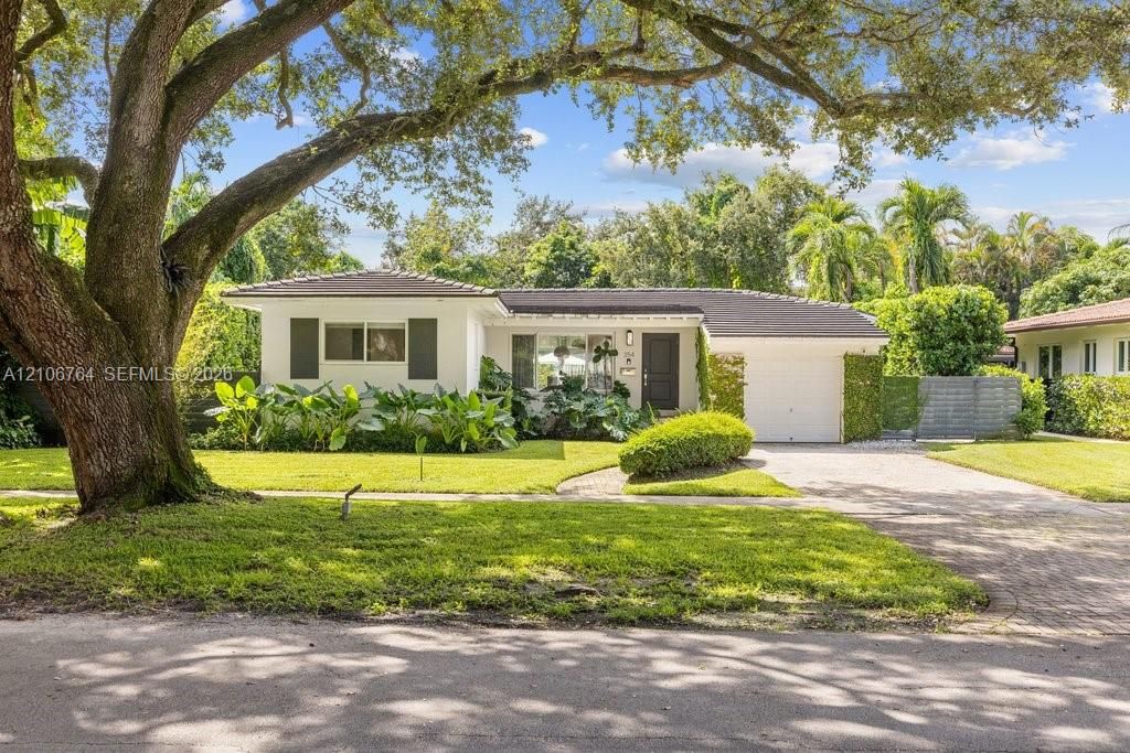 Single-story exterior featuring a light-toned stucco façade, dark-toned roof shingles, a paved driveway, and established landscape elements including a mature canopy tree
