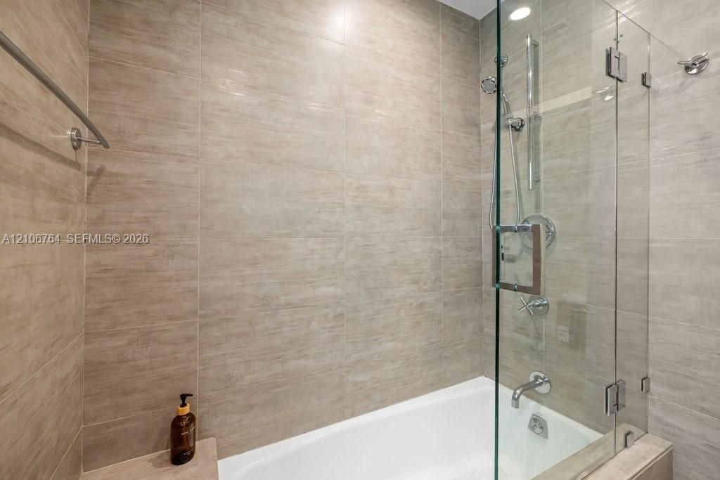 Full-height neutral-toned tile surround, frameless glass shower panel, chrome rainfall shower head with handheld attachment, built-in tub deck, and wall-mounted metal towel bar