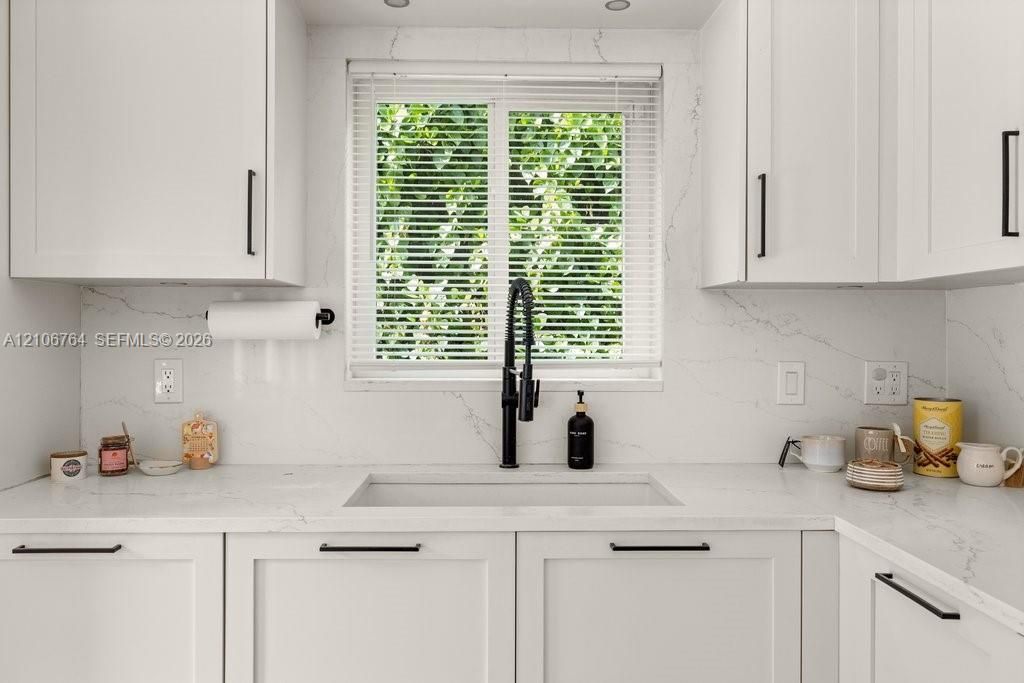 White shaker-style cabinetry with matte black hardware, stone-patterned countertops, integrated white apron-front sink, matte black gooseneck faucet, and full-height stone-patterned backsplash