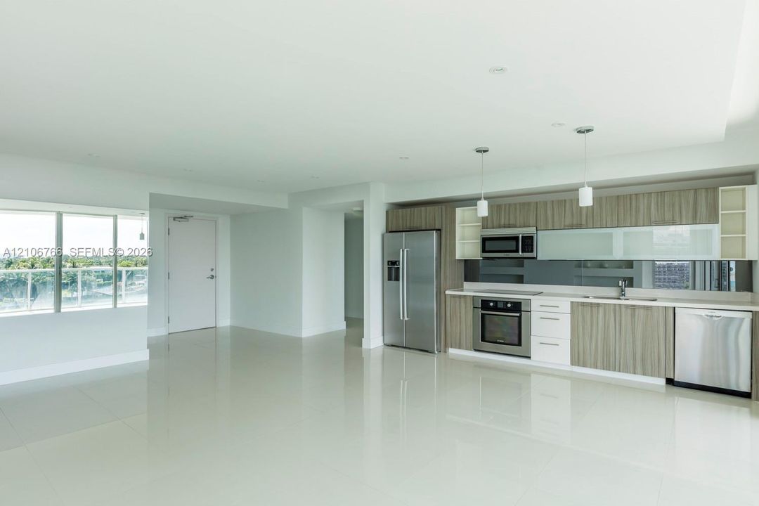 Open-concept area featuring polished tile flooring, wood-finish cabinetry, stainless steel appliances, glass-panel upper cabinet doors, and integrated pendant lighting