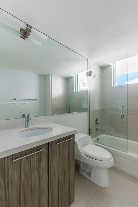 Bathroom featuring a wood-finish vanity with a stone-surface countertop, an integrated sink, a full-width wall mirror, and a glass-enclosed tub with tiled surround
