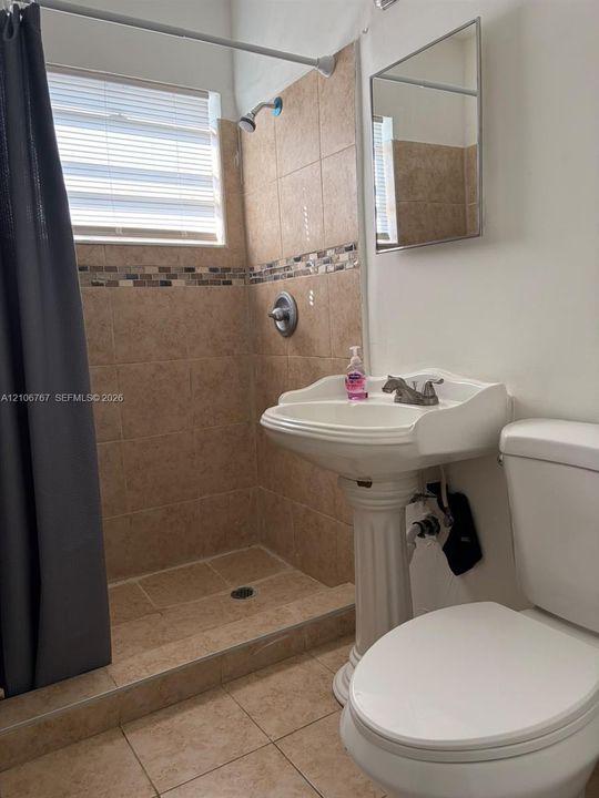 Bathroom featuring a pedestal sink, a walk-in shower with tile surround and decorative inlay, neutral floor tile, and a rectangular wall-mounted mirror