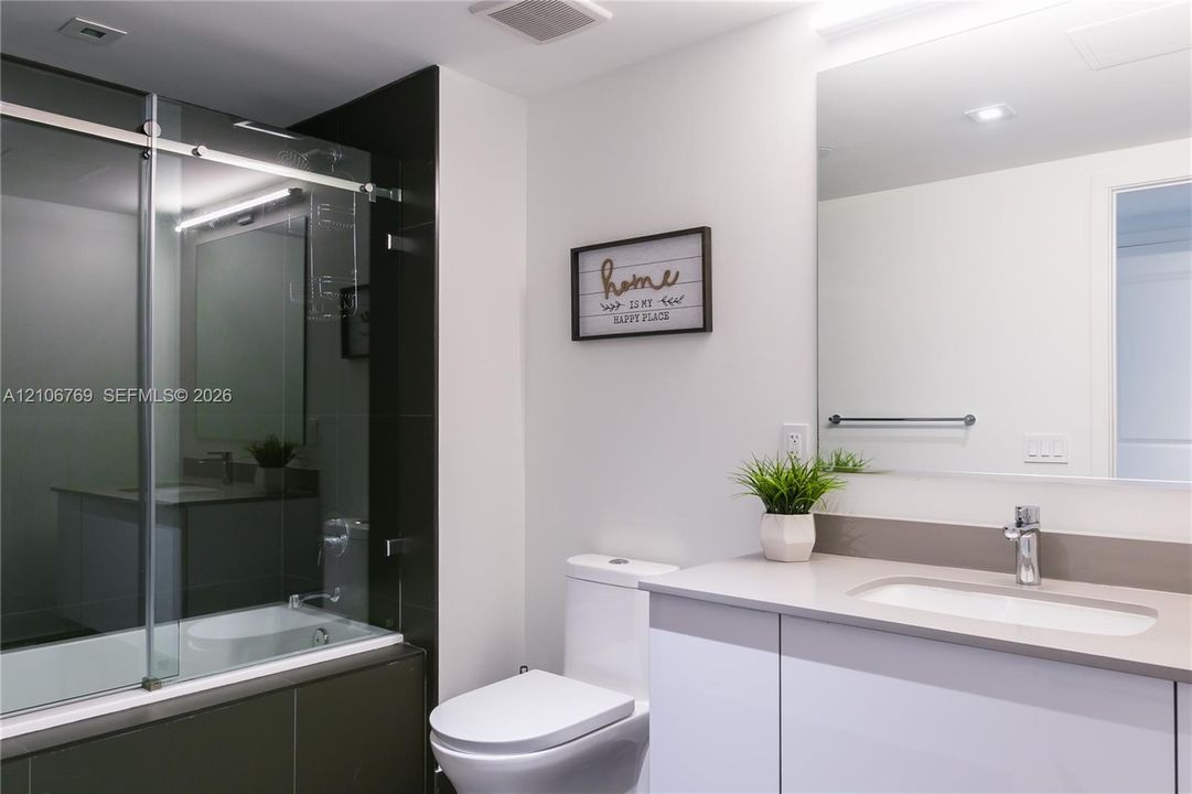 Full-width mirror, quartz countertop vanity with undermount sink, dual-flush toilet, and deep soaking tub enclosed by a sliding glass door with dark tile surround