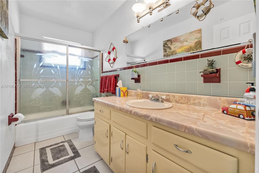 Bathroom featuring a stone-surface vanity with an integrated oval sink, ceramic tile wainscoting, a wall-to-wall mirror, and a tub-shower combination with sliding etched glass doors