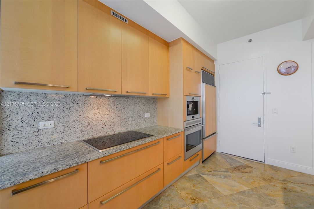 Kitchen featuring light-wood cabinetry with linear pulls, speckled stone countertops, a flush-mount electric cooktop, stainless steel wall-mounted oven, and stone tile flooring