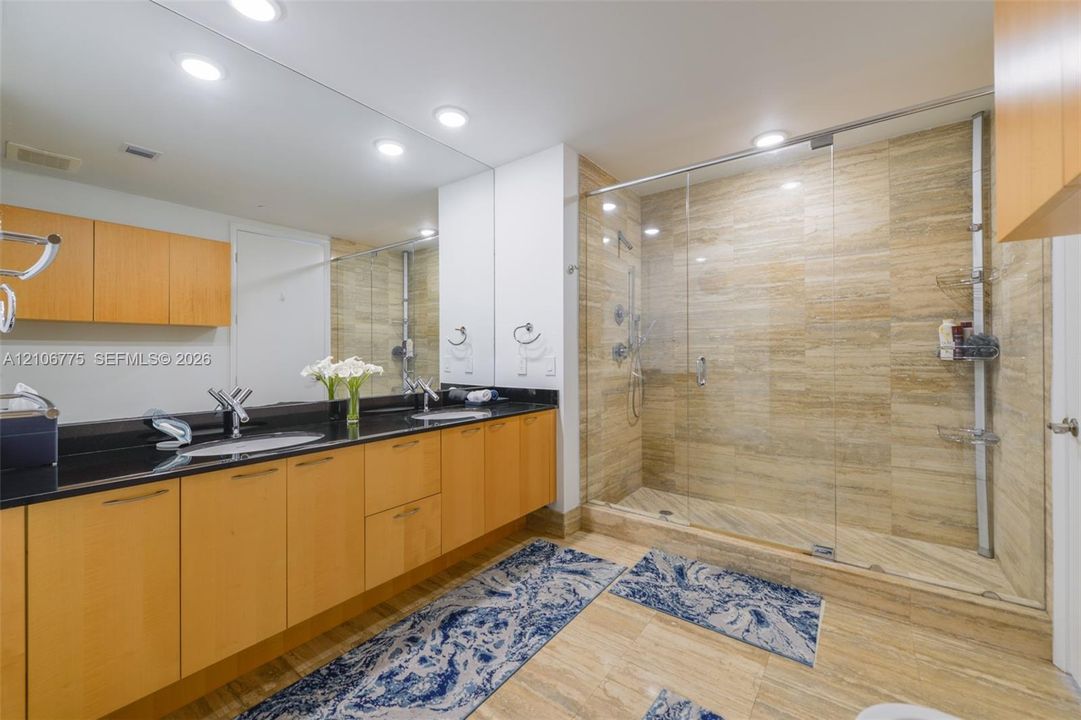 Full-width vanity with natural wood cabinetry and dark stone countertop, dual undermount sinks, expansive wall-to-wall mirror, and walk-in shower with floor-to-ceiling stone-tiling and frameless glass enclosure
