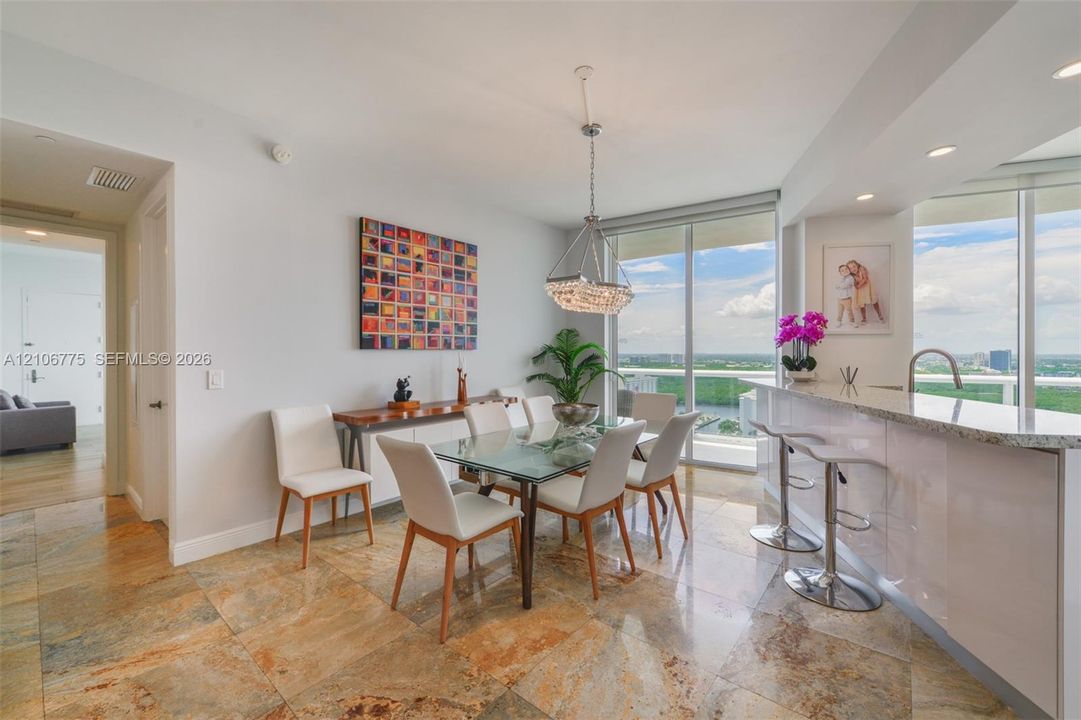 Open-concept dining area featuring oversized stone flooring, a tiered crystal chandelier, floor-to-ceiling windows, and an eat-in kitchen peninsula with a stone countertop