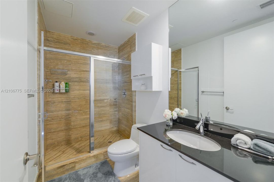 Bathroom featuring a walk-in shower with stone-tile surround, glass enclosure, polished countertop with undermount sink, and integrated cabinetry