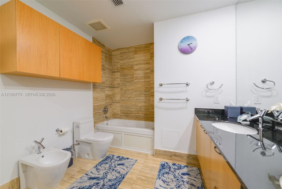 Bathroom featuring stone-finish wall and floor tiling, dark countertop with integrated sink, light wood-finish cabinetry, built-in tub with shower fixture, and wall-mounted bidet