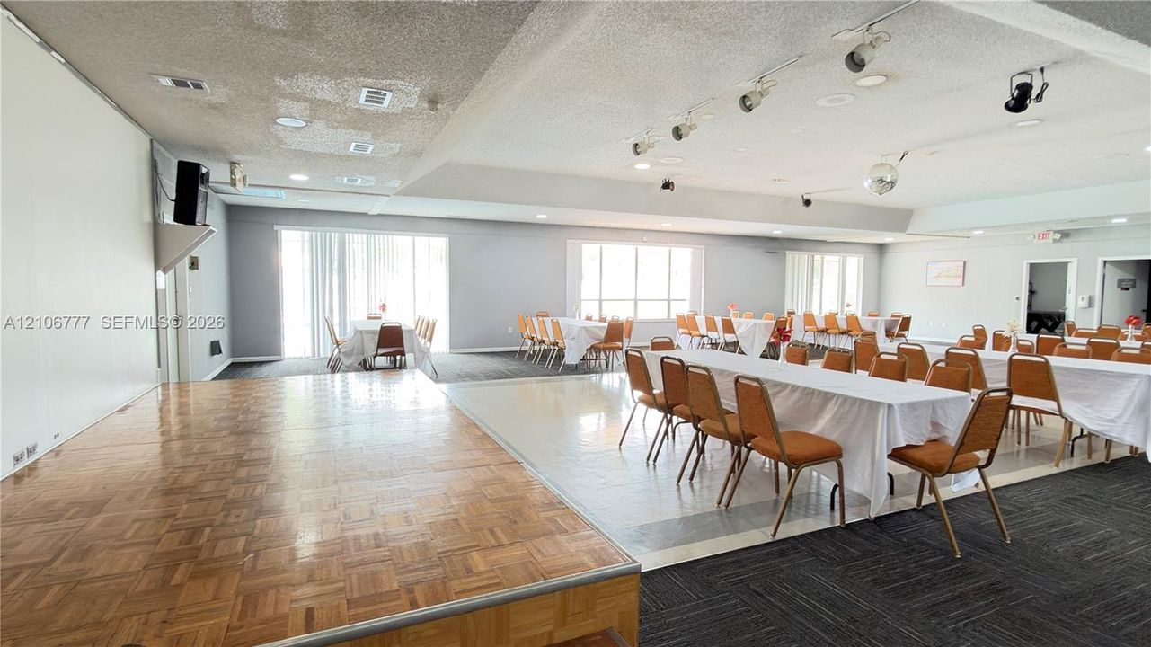 Expansive open-concept interior featuring a raised wood-parquetry platform stage, multi-section track lighting, large windows with vertical blinds, and a combination of tile and carpeted flooring