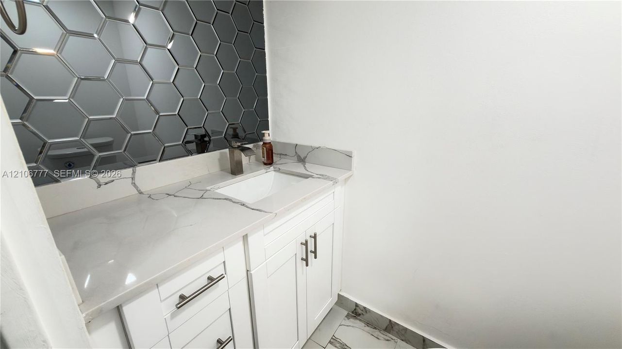 Vanity with white cabinetry, stone-surface countertop, integrated rectangular sink, metallic faucet, and decorative hexagonal mirror tiling