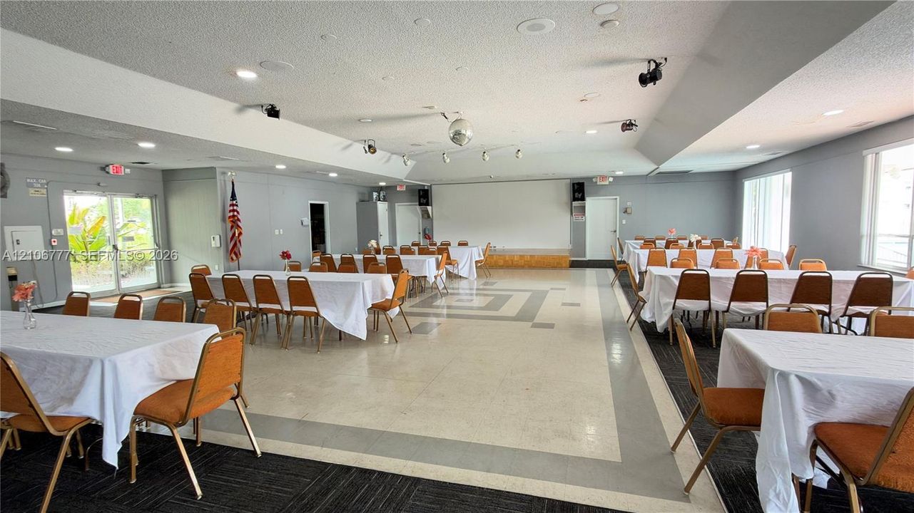 Expansive multi-purpose hall featuring a raised stage, recessed lighting, tile flooring with geometric border inlay, and dual-aspect glass-paneled exterior doors