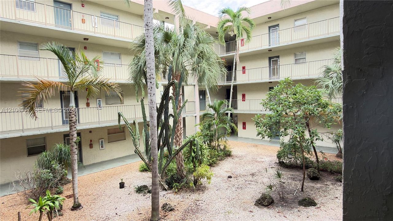 Multi-story building exterior featuring open-air walkways, metal balcony railings, decorative palm trees, and landscaped ground-level garden