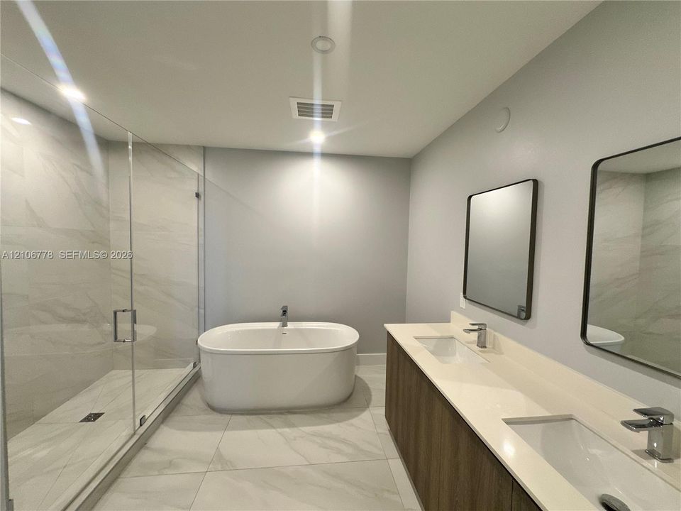 Double vanity with a composite countertop, wood-finish cabinetry, frameless walk-in glass shower, and freestanding soaking tub featuring large-format tile flooring