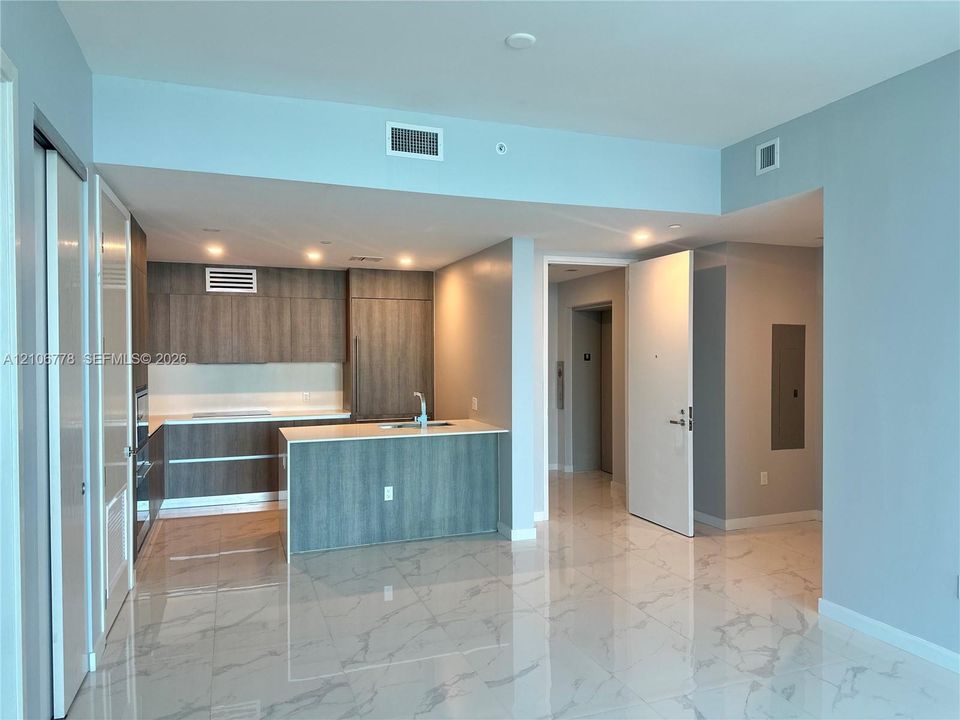 Modern open-concept living area featuring polished stone flooring, a central kitchen island with wood-finish cabinetry, stainless steel appliances, recessed ceiling lighting, and abundant natural light from expansive glass sliding doors