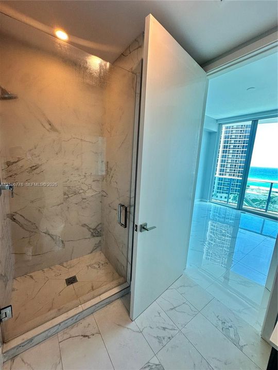 Walk-in shower featuring glass enclosure, ceiling-mounted fixture, and veined stone-finish wall tile, complemented by large-format polished flooring throughout the suite and floor-to-ceiling glass