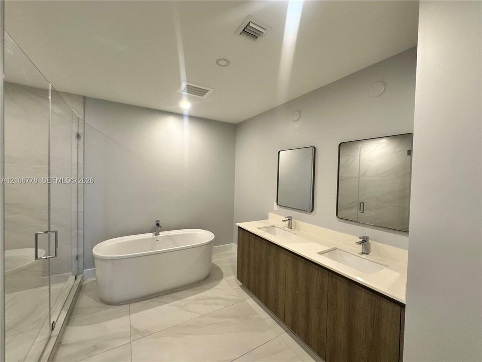 Dual-sink vanity with wood-finish cabinetry, quartz countertop, freestanding soaking tub, and glass-enclosed walk-in shower with tile surround