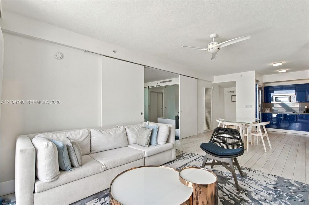 Open-concept interior featuring wood-finish flooring, sliding floor-to-ceiling mirror panels, a white ceiling fan, and high-gloss blue cabinetry in the kitchen