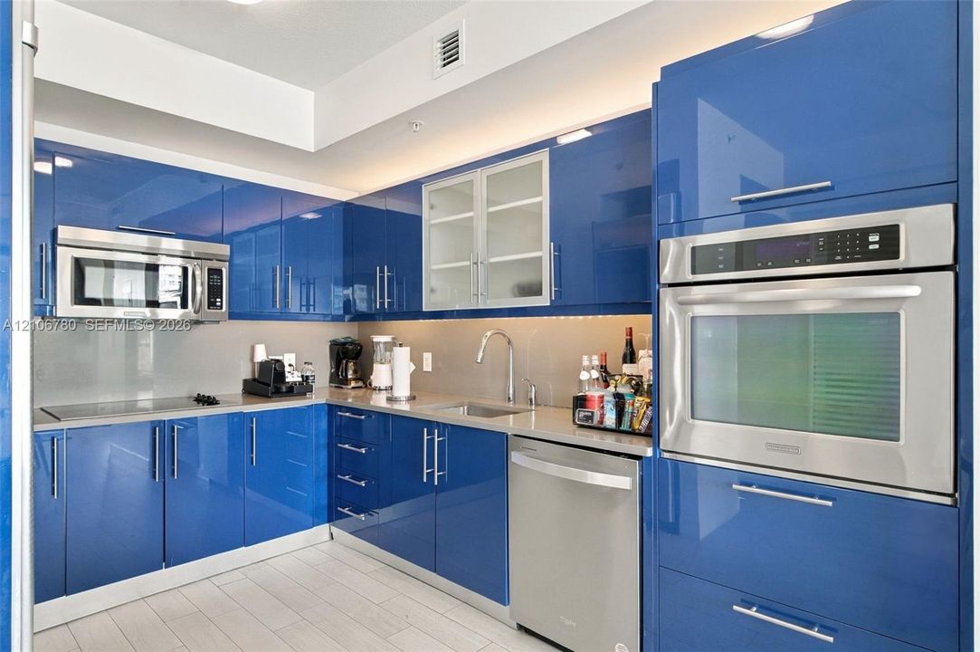 Glossy blue cabinetry, high-gloss flat-panel doors, stainless steel wall-mounted oven and microwave, neutral stone-finish countertops, and stainless steel undermount sink