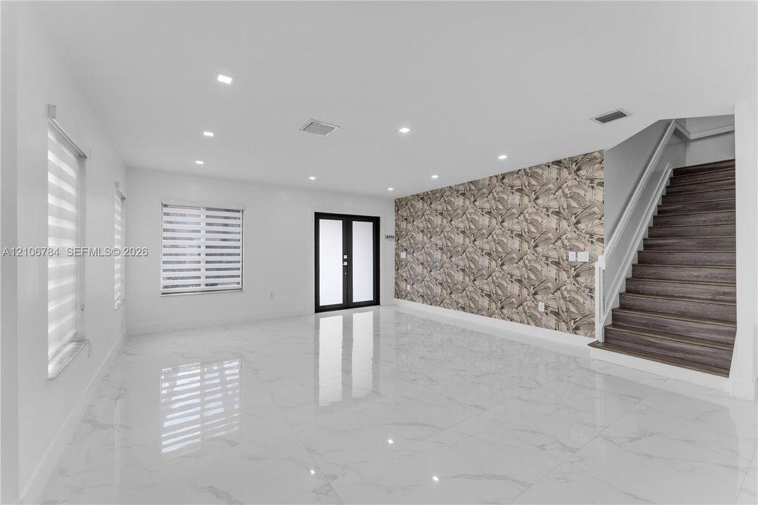 Open-concept room featuring large-format tile flooring, recessed lighting, double-door entryway, patterned accent wall, and staircase with wood-finish treads