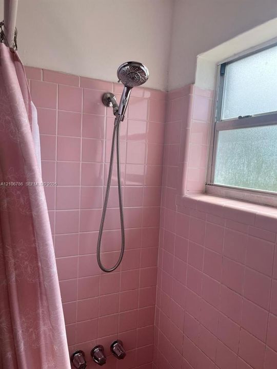 Shower enclosure featuring pink square ceramic wall tile, a handheld chrome shower head, and matching chrome plumbing fixtures, complemented by a recessed window with a sill