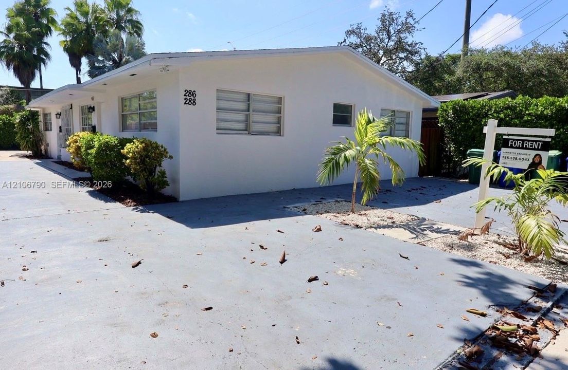 Single-story structure featuring a painted masonry exterior, horizontal louvered windows, and a paved concrete parking area