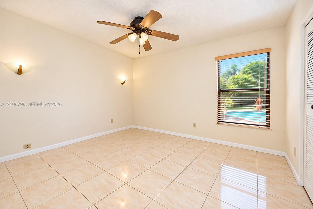 Neutral-toned tile flooring, ceiling fan with integrated light fixture, dual wall-mounted light sconces, louvered closet doors, and window with built-in blinds
