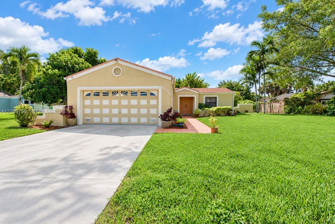 Single-story exterior featuring a stucco facade, attached two-car garage with arched decorative paneling, paved concrete driveway, and red brick walkway