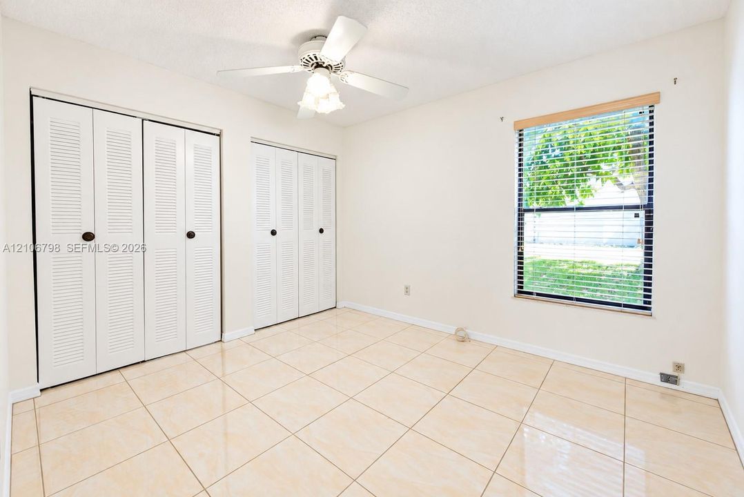 Interior room featuring large-format neutral tile flooring, dual louvered-style closets, a white ceiling fan with integrated light fixture, and a single window with wood-finish trim