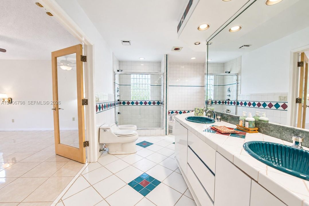Bathroom featuring a dual-sink vanity with tiled countertops, two ribbed teal vessel sinks, a glass-enclosed shower with glass block window, and white tile flooring with decorative accent insets