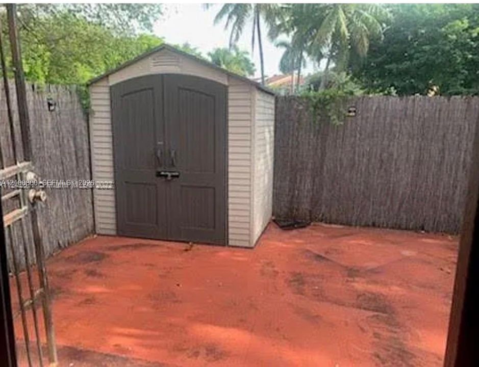 Exterior storage shed with double doors, set upon a red-toned concrete patio enclosed by reed fencing and an iron gate