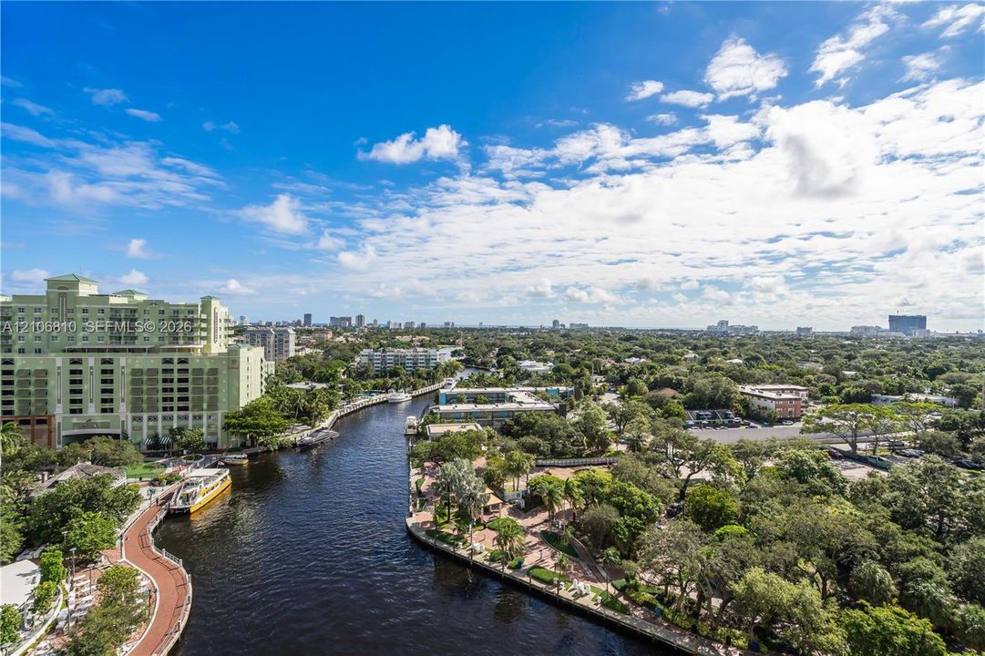 Elevated aerial perspective featuring a winding waterway, paved pedestrian paths, mature tropical landscaping, and a broad cityscape horizon