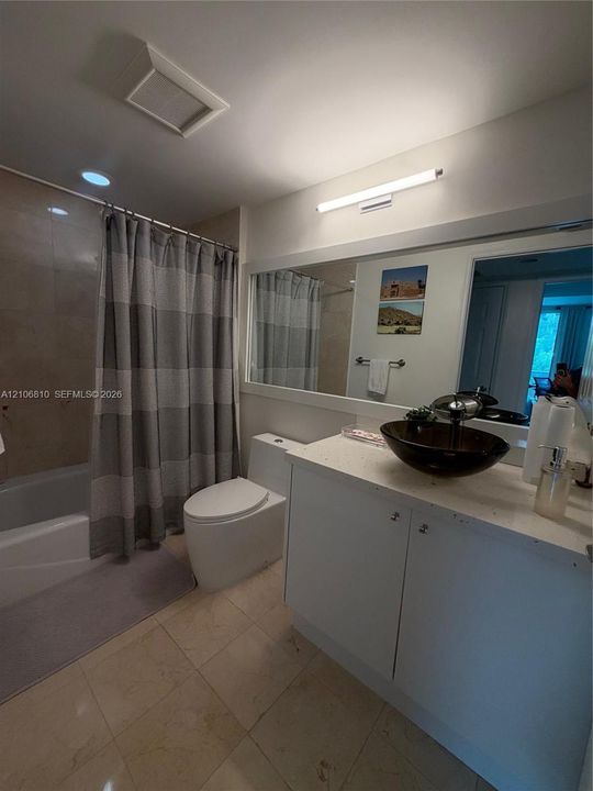Bathroom featuring stone-finish floor tiling, a white vanity with a light-colored solid surface countertop, a dark glass vessel sink, and a large wall-mounted mirror