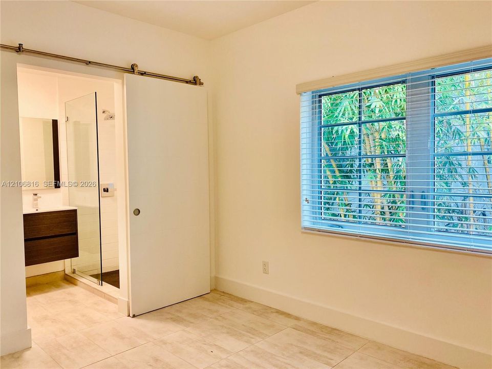 Interior space featuring tile flooring, sliding barn door with metal hardware, multi-pane window with horizontal blinds, and attached bathroom with floating vanity and glass-enclosed shower