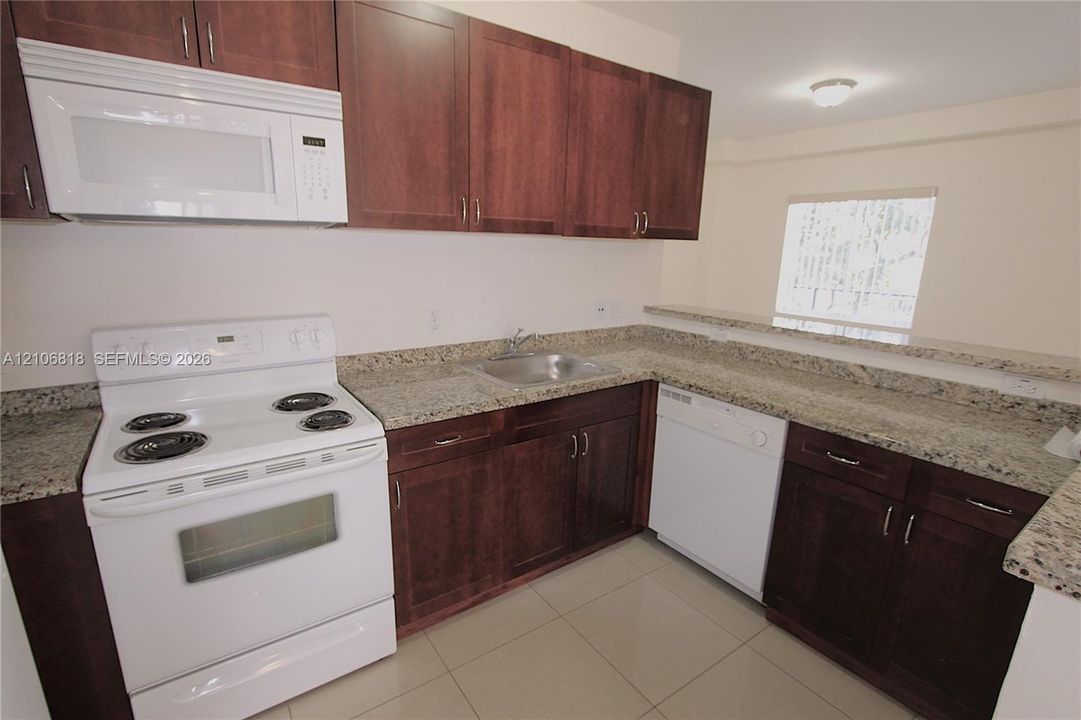 Kitchen featuring dark wood-finish cabinetry, stone-surface countertops, tile flooring, and white appliances including a built-in microwave, range, and dishwasher